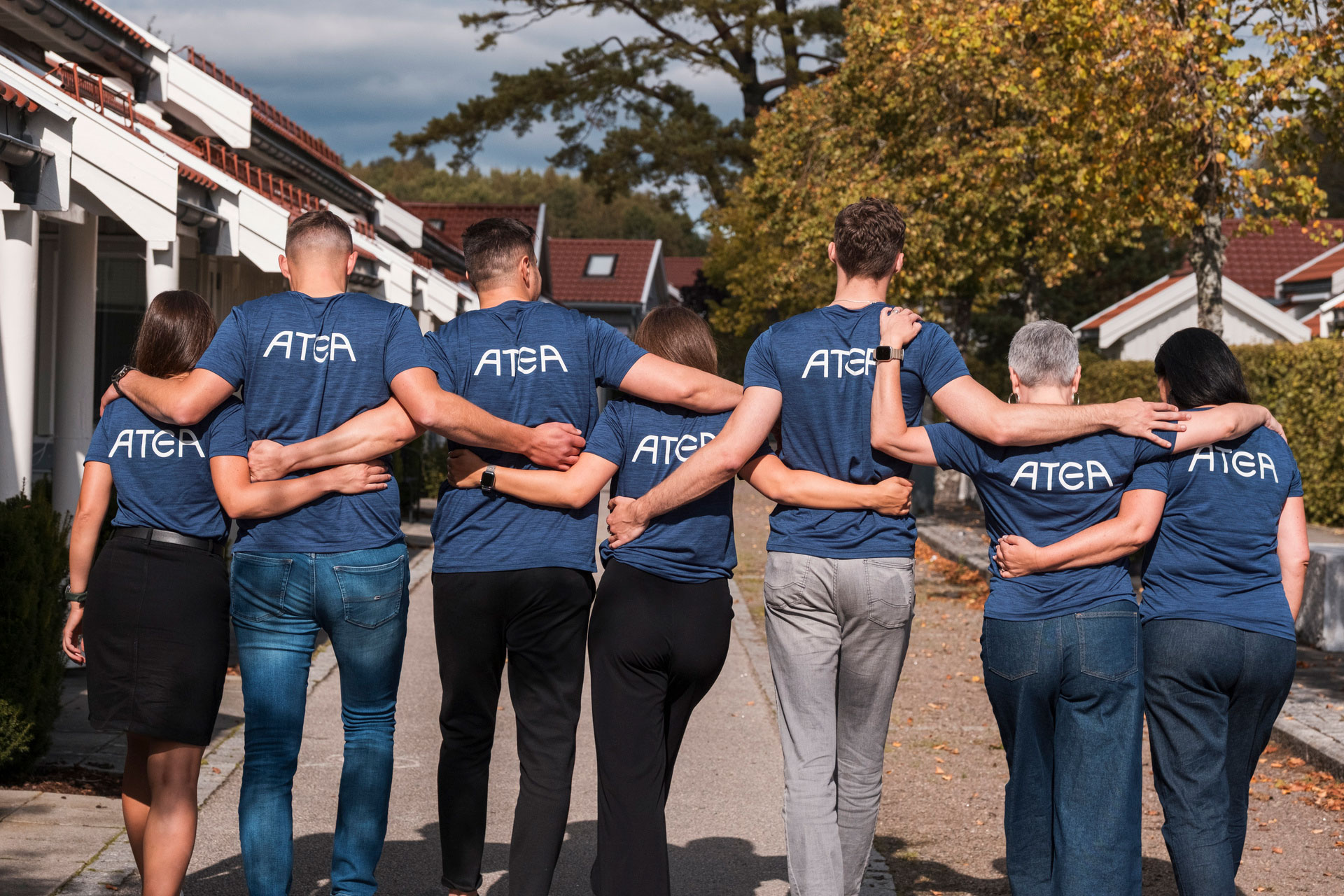 Seven People Walking With Their Backs Turned And Atea Logo On Their T Shirts